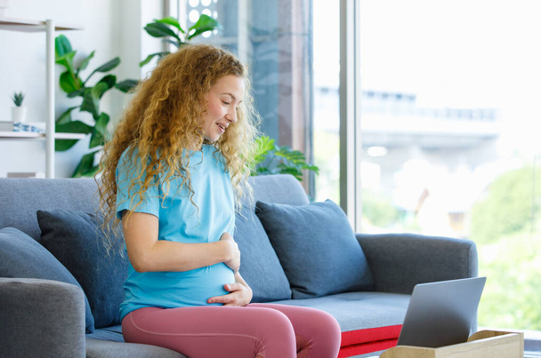 Caucasian happy pretty long curly hair healthy pregnancy mother in casual outfit sitting smiling on cozy sofa touching rubbing tummy belly watching online streaming via laptop computer in living room. - Foto, Imagen