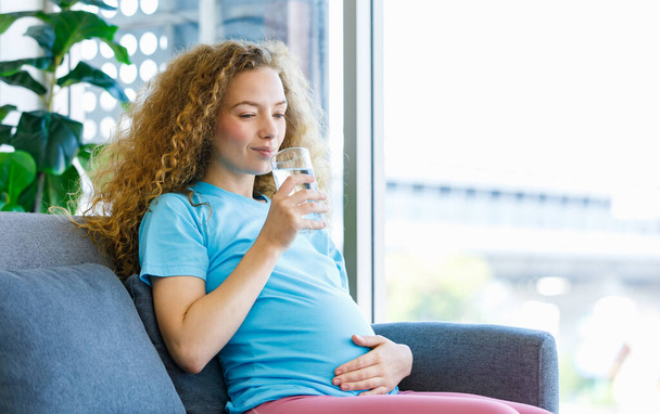Caucasian happy pretty long curly hair healthy thirsty pregnancy mother in casual outfit sitting smiling on cozy sofa drinking water from glass holding rubbing big belly tummy expecting for newborn. - Foto, Bild