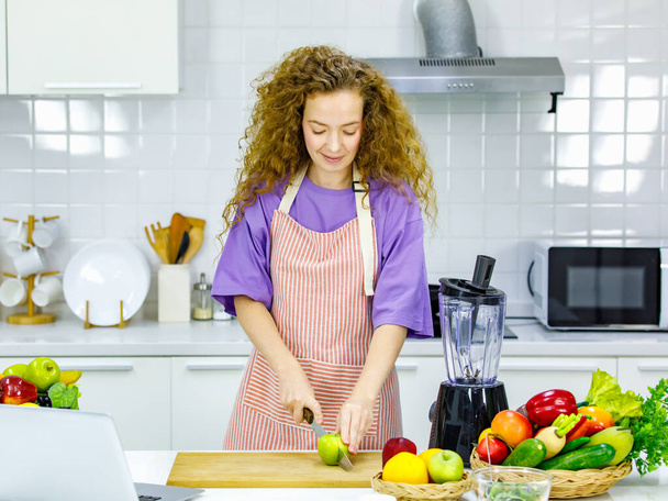 Caucasian happy pretty curly hair healthy big belly tummy pregnancy mother in apron standing learning cooking online from laptop computer holding knife cutting vegetable on wooden board in kitchen. - Foto, Imagen