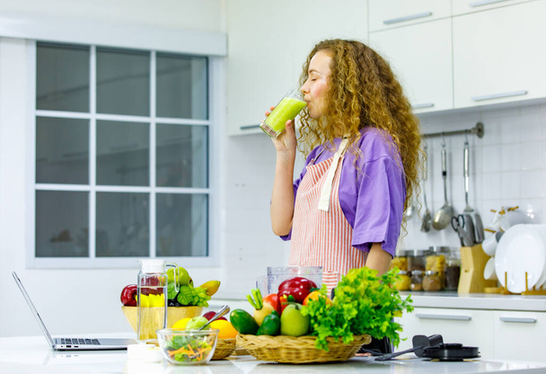 Caucasian happy curly hair healthy big belly tummy pregnancy mother in apron standing smiling learning online cooking via laptop drinking organic blending mixed vegetable and fruit juice in kitchen. - 写真・画像