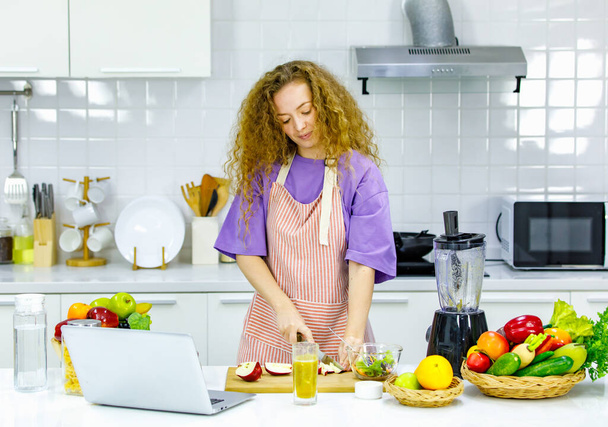 Καυκάσιος χαρούμενος σγουρά μαλλιά υγιή μεγάλη κοιλιά εγκυμοσύνη μητέρα σε ποδιά στέκεται εκμάθηση μαγειρικής σε απευθείας σύνδεση από τον υπολογιστή laptop κρατώντας μαχαίρι αποφλοίωση κόκκινο μήλο στο ξύλινο διοικητικό συμβούλιο στην κουζίνα. - Φωτογραφία, εικόνα