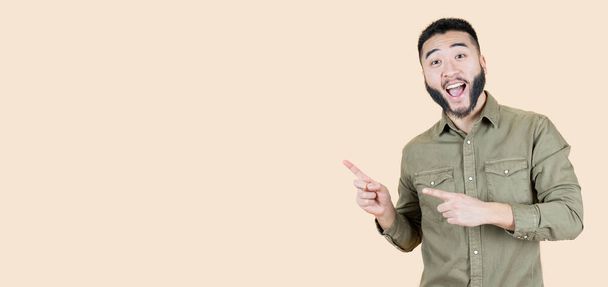 handsome young asian man, smiling and looking at the camera pointing with two hands and fingers to the side. beige background - Photo, Image