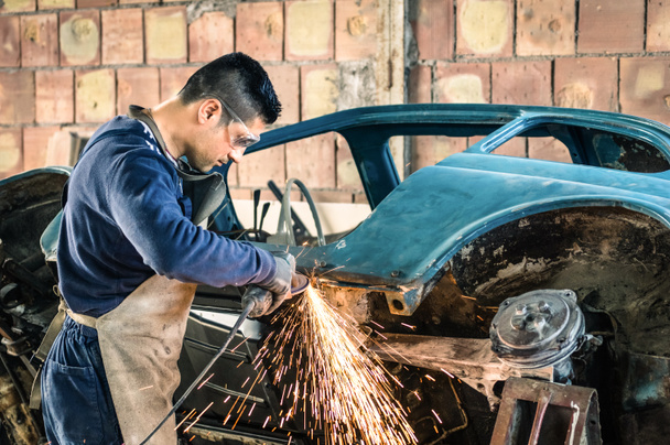Nuori mies mekaaninen työntekijä korjaamassa vanhaa vintage-auton runkoa sotkuisessa autotallissa - Työturvallisuus suojavaatetuksella
 - Valokuva, kuva
