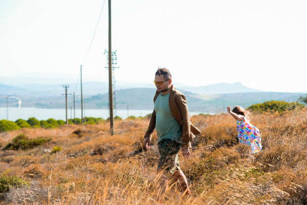 Apa és lánya sétál a gyönyörű mezők hegyek a háttérben. Szélmalom mező. Elektromos rudak. A megújuló energia fogalma, a természet, a család, az elektromosság, a zöld, a jövő szeretete - Fotó, kép
