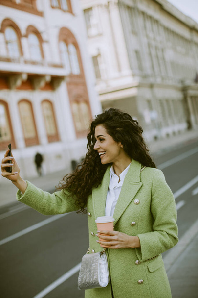 Pretty young woman using smartphone to take selfie photo on the street and holding takeaway coffee - Foto, Imagem