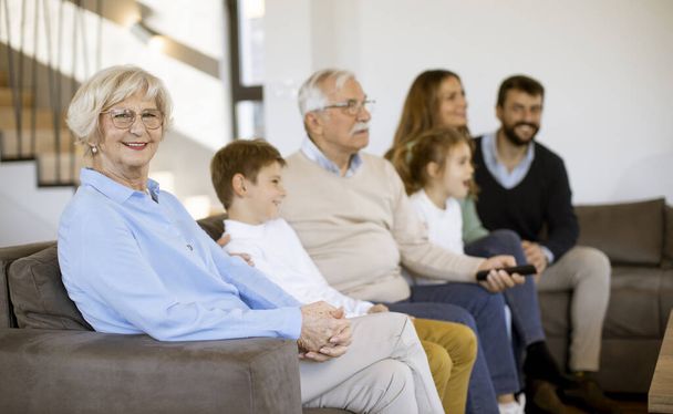 Wielopokoleniowa rodzina siedzi razem na kanapie w nowoczesnym domu i ogląda telewizję - Zdjęcie, obraz