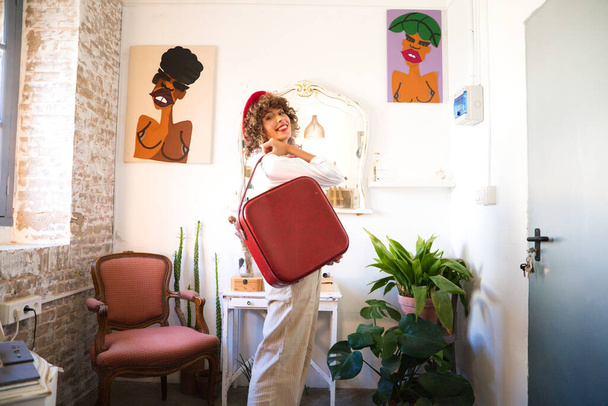 Молодая, красивая, богемная латиноамериканка с красным беретом на голове и красным винтажным чемоданом, счастливо смотрящая в камеру для фотографии. Концепция предприимчивой женщины. - Фото, изображение
