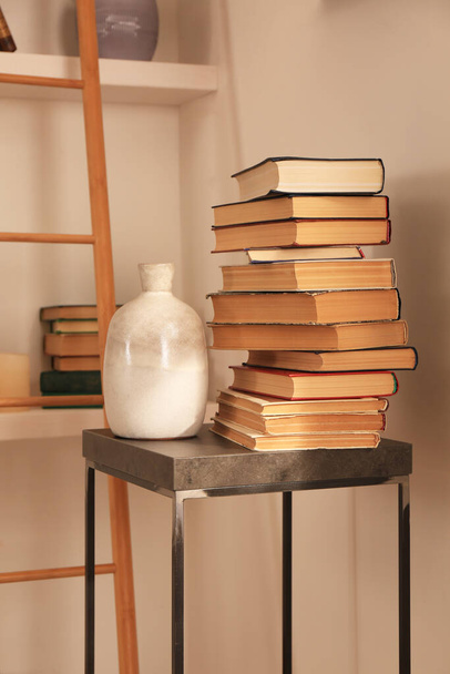 Stack of different books and vase on table near bookshelves in home library - Φωτογραφία, εικόνα
