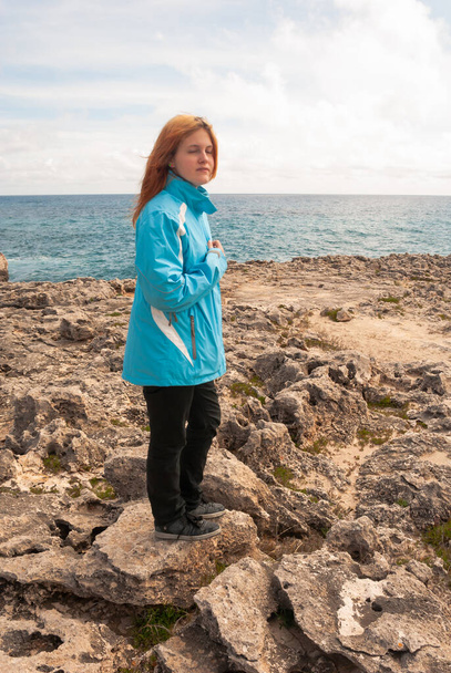Young woman with orange hair, wearing a blue jacket, standing on a rock in front of the ocean, meditating with eyes closed, holding her hand on her chest, majorca - Photo, Image
