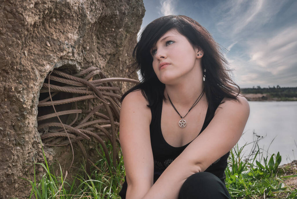 Молодая готическая эмо панк-девушка сидит у озера, черные волосы, в черной одежде, смотрит вверх, горизонтально - Фото, изображение