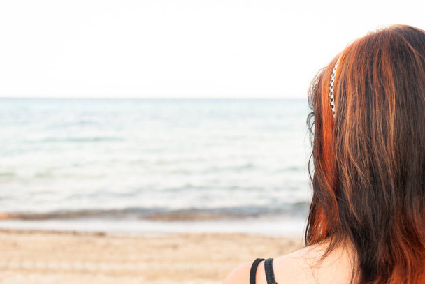 Kızıl ve siyah uzun saçlı genç bir kadın sahilde dikiz aynasından denize bakıyor. - Fotoğraf, Görsel