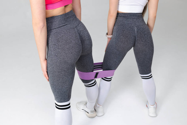 Two women in beautiful fitness leggings with attractive buttocks and a fitness elastic band pose in the studio - Photo, Image