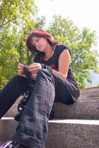 Chica emo punk, adulto joven con pelo rojo negro, sentado en una escalera al aire libre, mirando hacia abajo, vista de ángulo bajo, vertical - Foto, Imagen