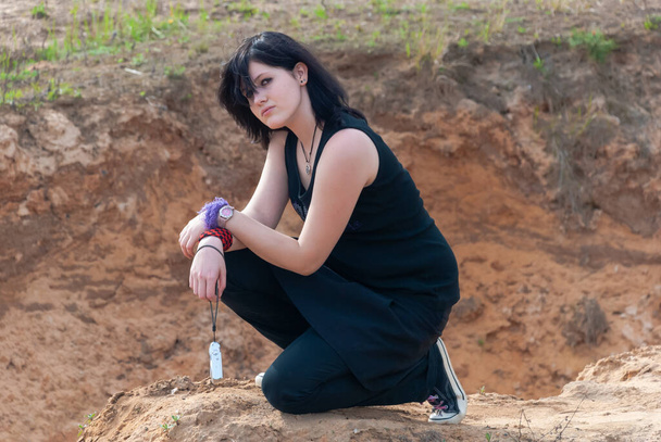 Punk emo meisje, jong volwassene met lang zwart haar, knielen op een heuvel buiten in de natuur, met een camera in haar linkerhand, kijken naar de camera, horizontaal - Foto, afbeelding