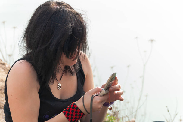 Punk emo girl with long black hair and eyeliner, young adult using her mobile phone while sitting outdoors at the lake, holding a camera in her right hand, horizontal - Photo, Image