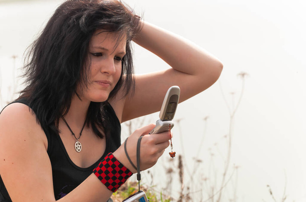 Punk emo girl with long black hair and eyeliner, young adult using her mobile phone while sitting outdoors at the lake, holding a camera in her right hand, horizontal - Photo, Image