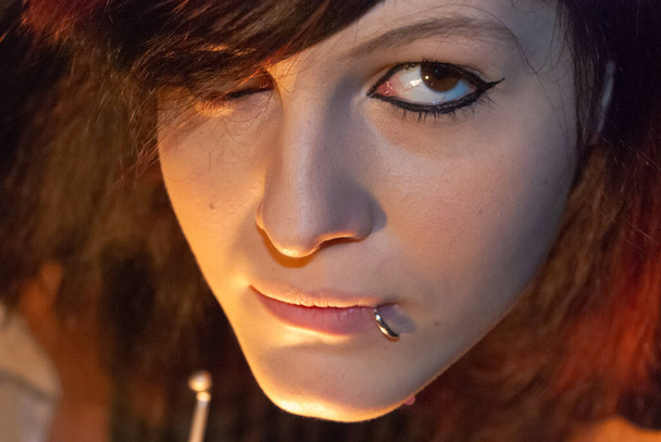 Νεαρό πανκ emo κορίτσι με σκουλαρίκια, με κόκκινα και μαύρα μαλλιά, κοιτάζοντας κάμερα με σοβαρή έκφραση του προσώπου, κοντινό πλάνο - Φωτογραφία, εικόνα