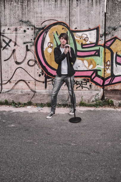 Молодой взрослый певец, с каштановыми длинными волосами и выступающей бородой, рок-музыкант с микрофоном на улице перед стеной с граффити, стоящий и смотрящий на камеру, одетый в джинсы и футболку - Фото, изображение