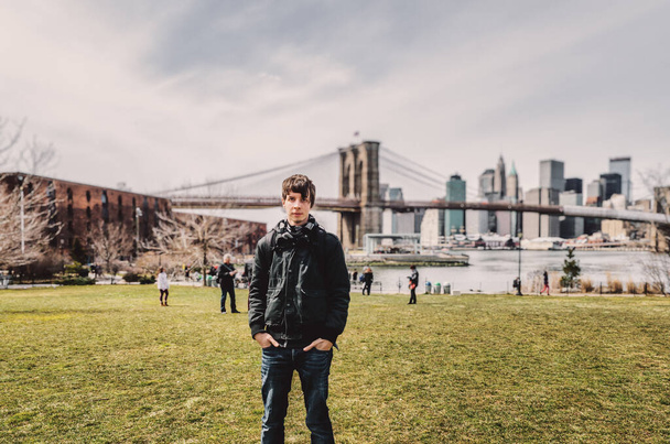 ブルックリンに立っている若い大人の男は、ニューヨークのマンハッタンの街並みを背景にカメラ、ブルックリン橋、スカイラインを見ている牧草地、ニューヨーク、ニューヨーク、アメリカ - 写真・画像