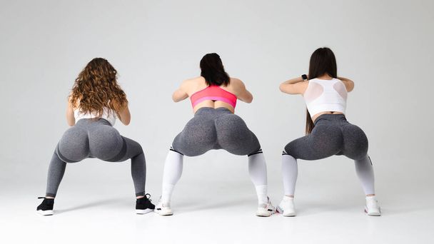 Three young fitness women squat in the studio. Girls with beautiful buttocks are doing a warm-up - 写真・画像