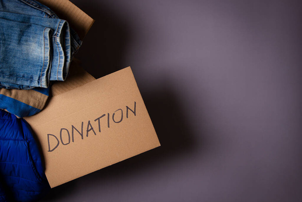 Caja de cartón con ropa y tarjeta con inscripción Donación sobre fondo gris. Ropa para donación, caridad. Vista superior, espacio de copia - Foto, Imagen