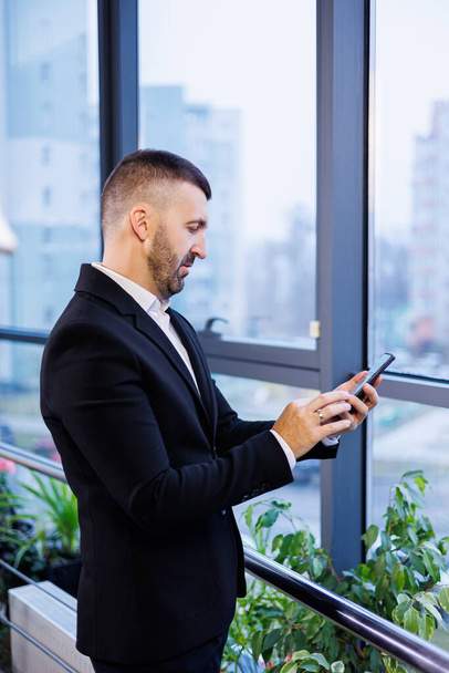 Portrét stylového mladého podnikatele stojícího ve velké moderní kanceláři vysoko v nejvyšším patře a dívajícího se na telefon - Fotografie, Obrázek