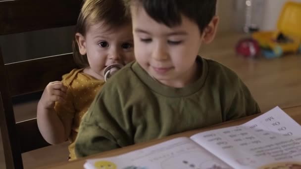 Der Junge sitzt mit seiner kleinen Schwester auf dem Stuhl vor dem Tisch, an dem er seine Hausaufgaben gemacht hat, während seine Mutter das Notizbuch des Jungen schließt, der - Filmmaterial, Video