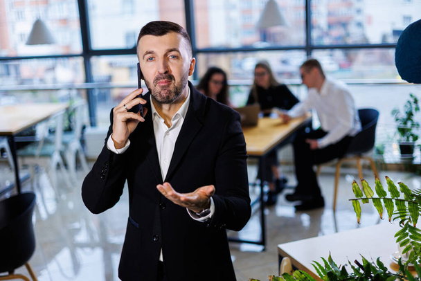 Όμορφος νέος επιχειρηματίας με μοντέρνα στολή, χρησιμοποιεί το κινητό του τηλέφωνο, πετυχημένος άντρας μιλάει στο τηλέφωνο στο γραφείο. - Φωτογραφία, εικόνα