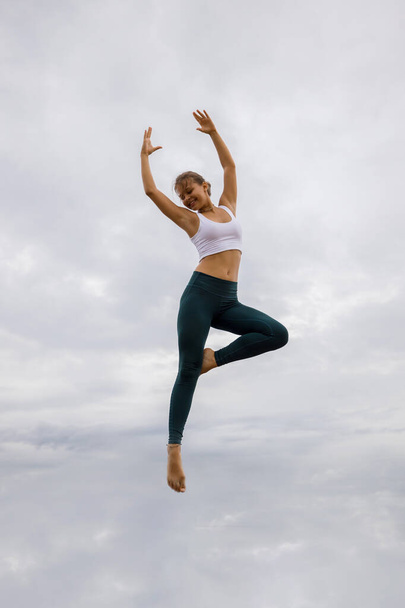 Όμορφη νεαρή γυναίκα πηδάει πάνω από συννεφιασμένο ουρανό. Λευκή γυναίκα που φοράει αθλητικά. Γυμναστική, έννοια ευεξίας. Εξωτερική δραστηριότητα. Αντιγραφή χώρου. Ουράνιο φόντο. Μπαλί, Ινδονησία - Φωτογραφία, εικόνα