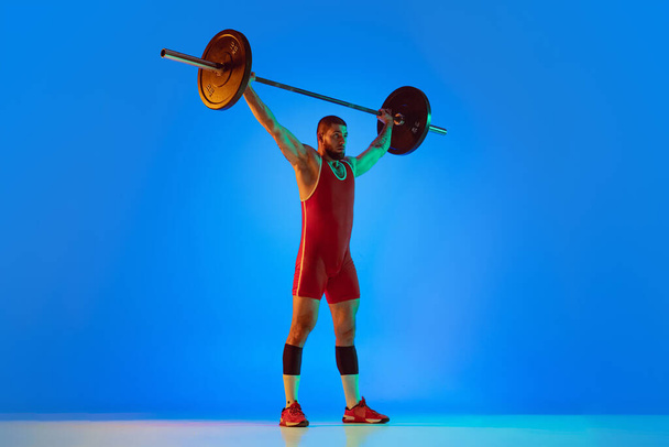 Студийный снимок молодого человека в красной спортивной одежде, упражняющегося с барбекю изолированным синим фоном в неоне. Sport, heavy tlifting, power, achievements concept - Фото, изображение