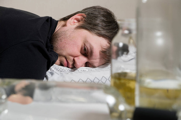 Ένας μεθυσμένος αλκοολικός ξαπλώνει στο κρεβάτι, με άδεια μπουκάλια στο τραπέζι. Αλκοολισμός, εξάρτηση από το αλκοόλ ως κοινωνικό πρόβλημα - Φωτογραφία, εικόνα