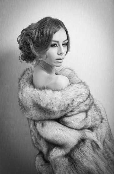 Attractive sexy young woman wearing a fur coat posing provocatively indoor. Portrait of sensual female with creative haircut, studio shot. Beautiful girl covered only with a fur exposing her shoulders - Фото, зображення