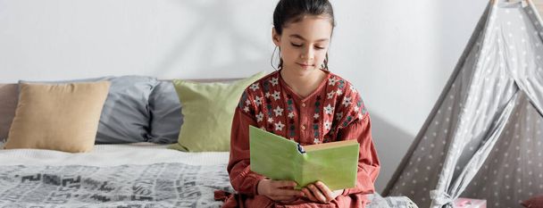preadolescente niña leyendo libro mientras está sentado en la cama cerca de almohadas y wigwam, pancarta - Foto, imagen