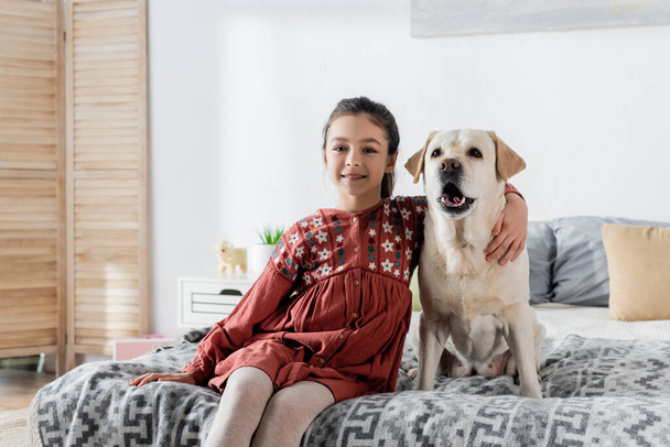 happy brunette girl embracing labrador and smiling at camera while sitting on bed - Fotografie, Obrázek