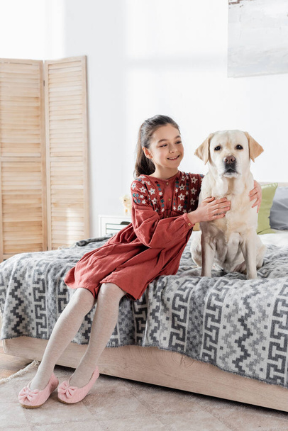 happy preteen girl cuddling labrador dog while sitting on bed at home - Foto, Bild