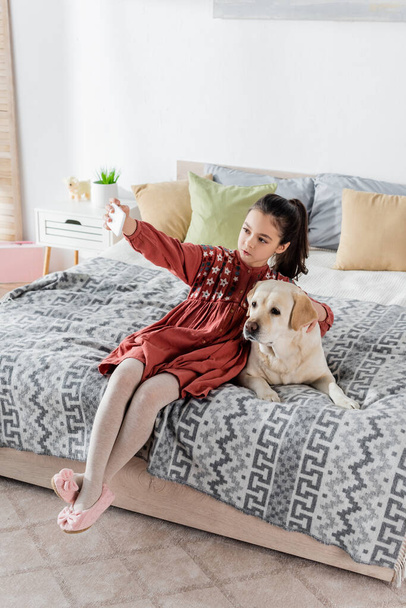 full length view of preteen girl taking selfie with labrador on bed at home - 写真・画像