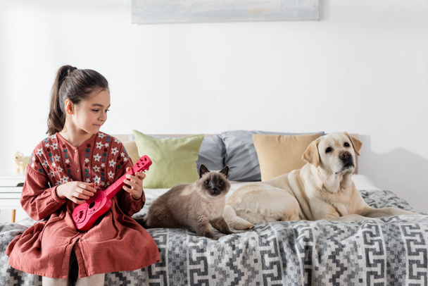 smiling girl playing toy guitar near cat and labrador dog on bed - Foto, afbeelding