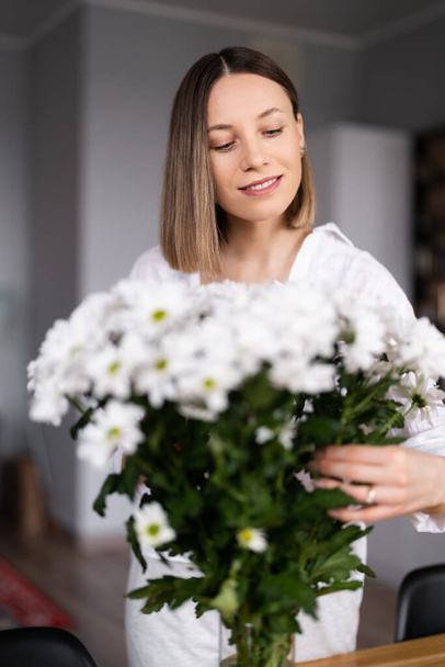 Beautiful woman putting fresh white flowers into a vase - Фото, изображение