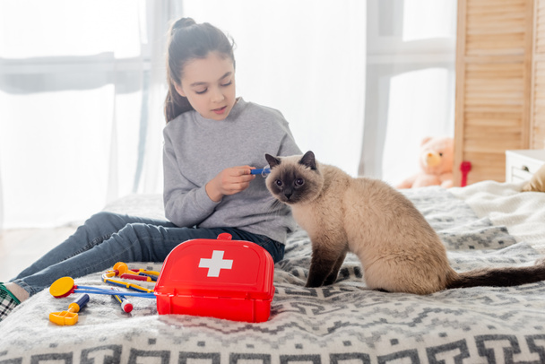 девушка делает инъекцию коту с игрушечным шприцем рядом с аптечкой на кровати - Фото, изображение