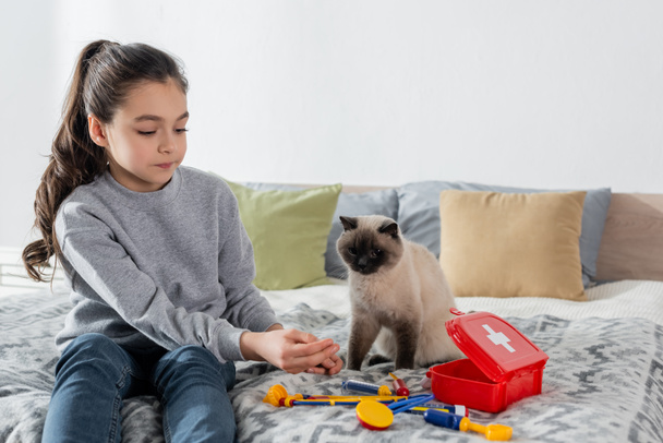 tyttö istuu sängyllä ja leikkii lääkäriä lelu lääketieteen asetettu lähelle kissa - Valokuva, kuva