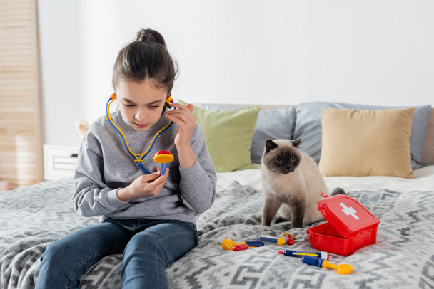 chica con juguete estetoscopio jugando doctor en cama cerca gato y botiquín de primeros auxilios - Foto, Imagen