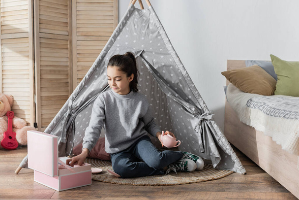 girl holding teapot while sitting in wigwam near box with toy tea set - Foto, Imagen