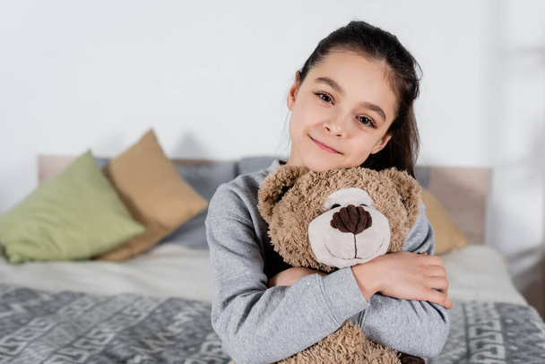 cheerful preteen girl embracing teddy bear and looking at camera in bedroom - Fotografie, Obrázek