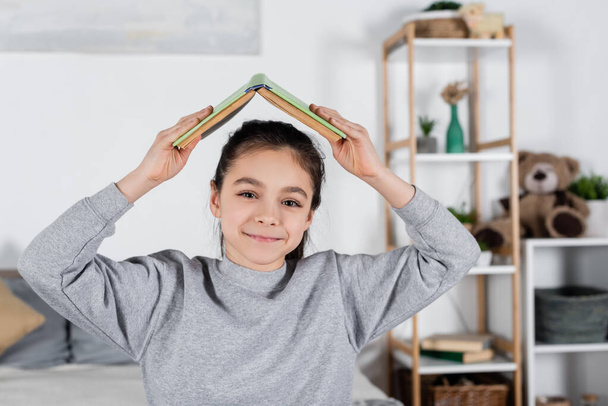 cheerful kid holding book above head and smiling at camera - Photo, image