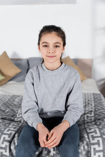 preteen girl smiling at camera while sitting on blurred bed - Zdjęcie, obraz
