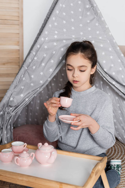 ragazza che beve tè dalla tazza giocattolo mentre gioca in wigwam - Foto, immagini