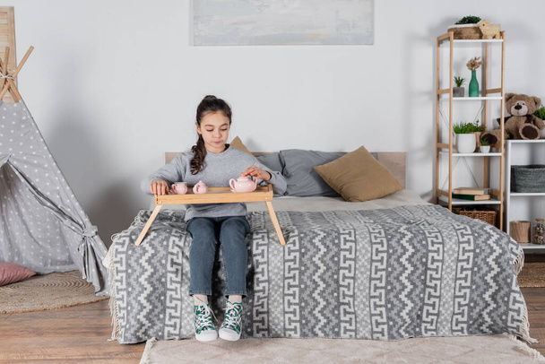 full length view of girl sitting on bed with toy tea set on wooden tray - Foto, Imagen