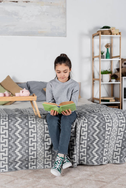 full length view of girl sitting on bed near tray with toy tea set and reading book - Photo, Image