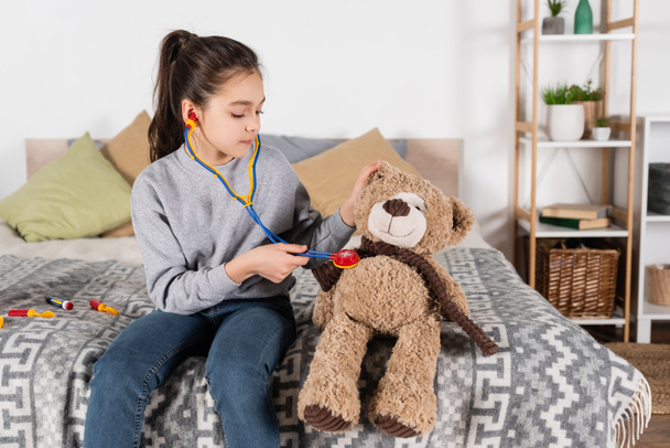 брюнетка девочка-подросток играет дома и рассматривает плюшевого мишку с помощью игрушечного стетоскопа - Фото, изображение