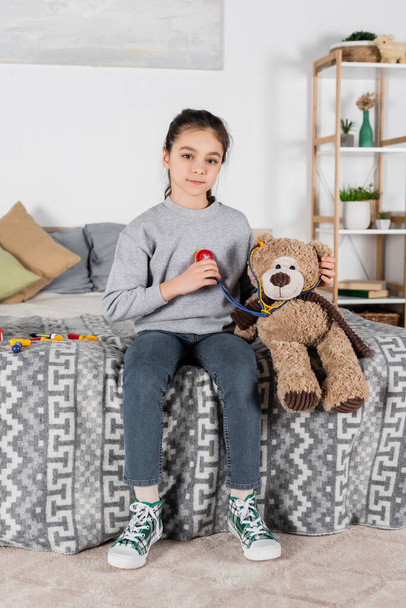 girl looking at camera while playing with teddy bear and toy stethoscope in bedroom - Photo, Image
